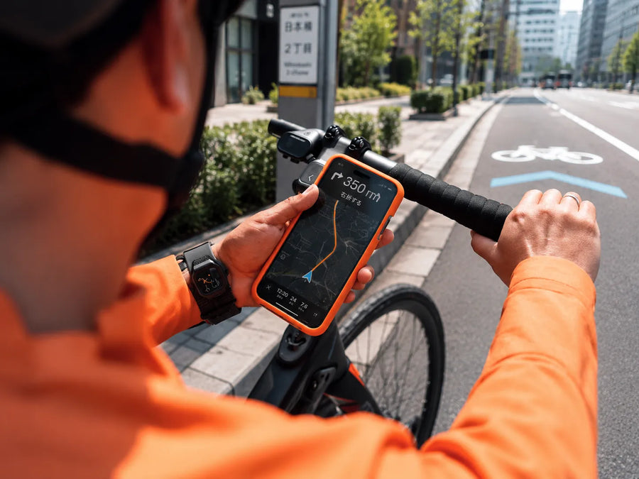 Torrasのオレンジケースのスマホを自転車ハンドルに固定し、ナビ画面を確認するヘルメット姿の男性。街路を進むサイクリングの様子。