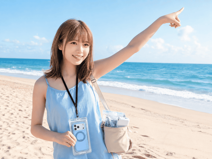 晴れた砂浜でTorrasの防水スマホケースを首に掛け遠くを指差す青い服を着た笑顔の女性