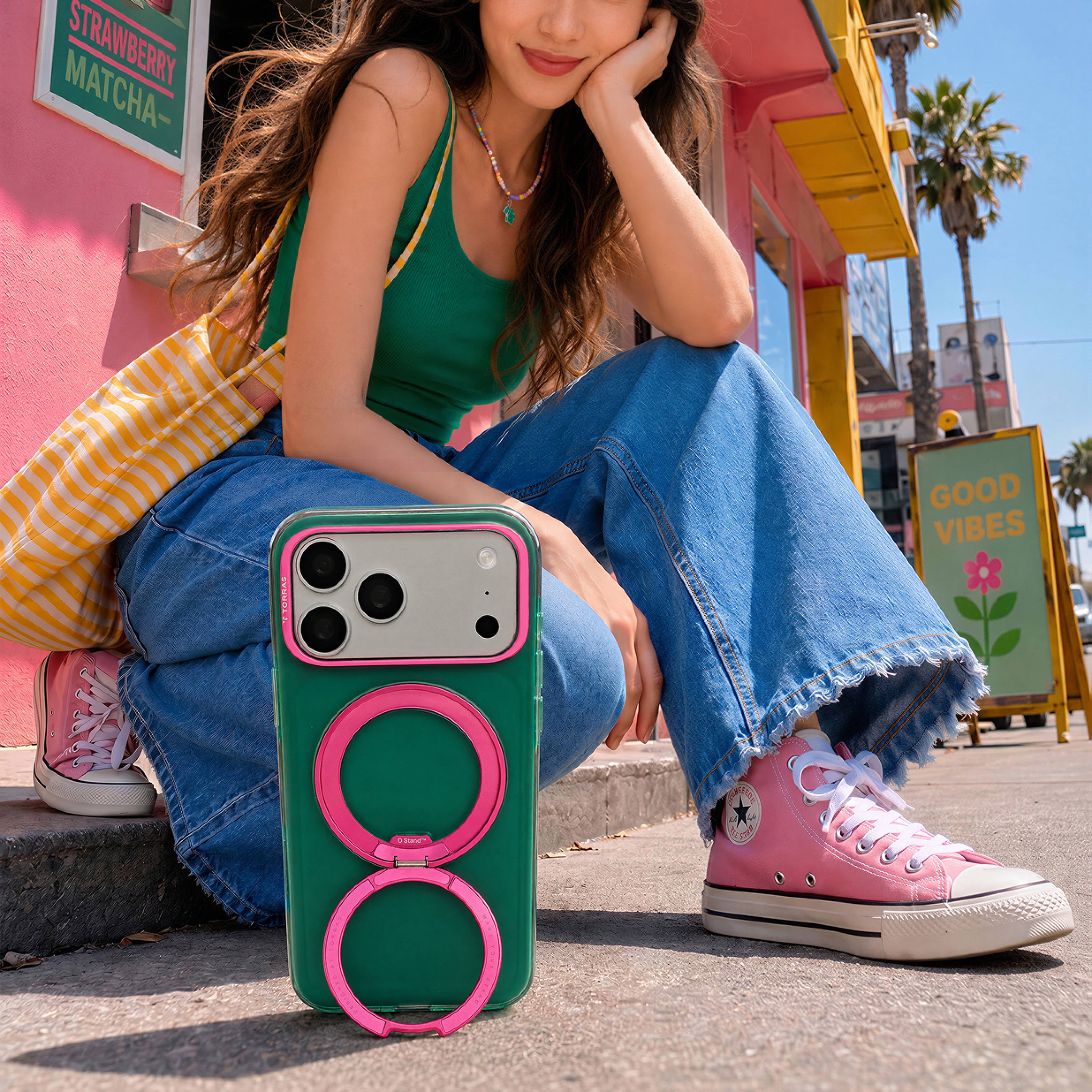TORRAS Ostand O3 Hue ケース iPhone 18 Pro/Pro Max 対応、屋外のベンチに座る女性、グリーンとピンクのケースが膝元に見えるライフスタイルシーン。